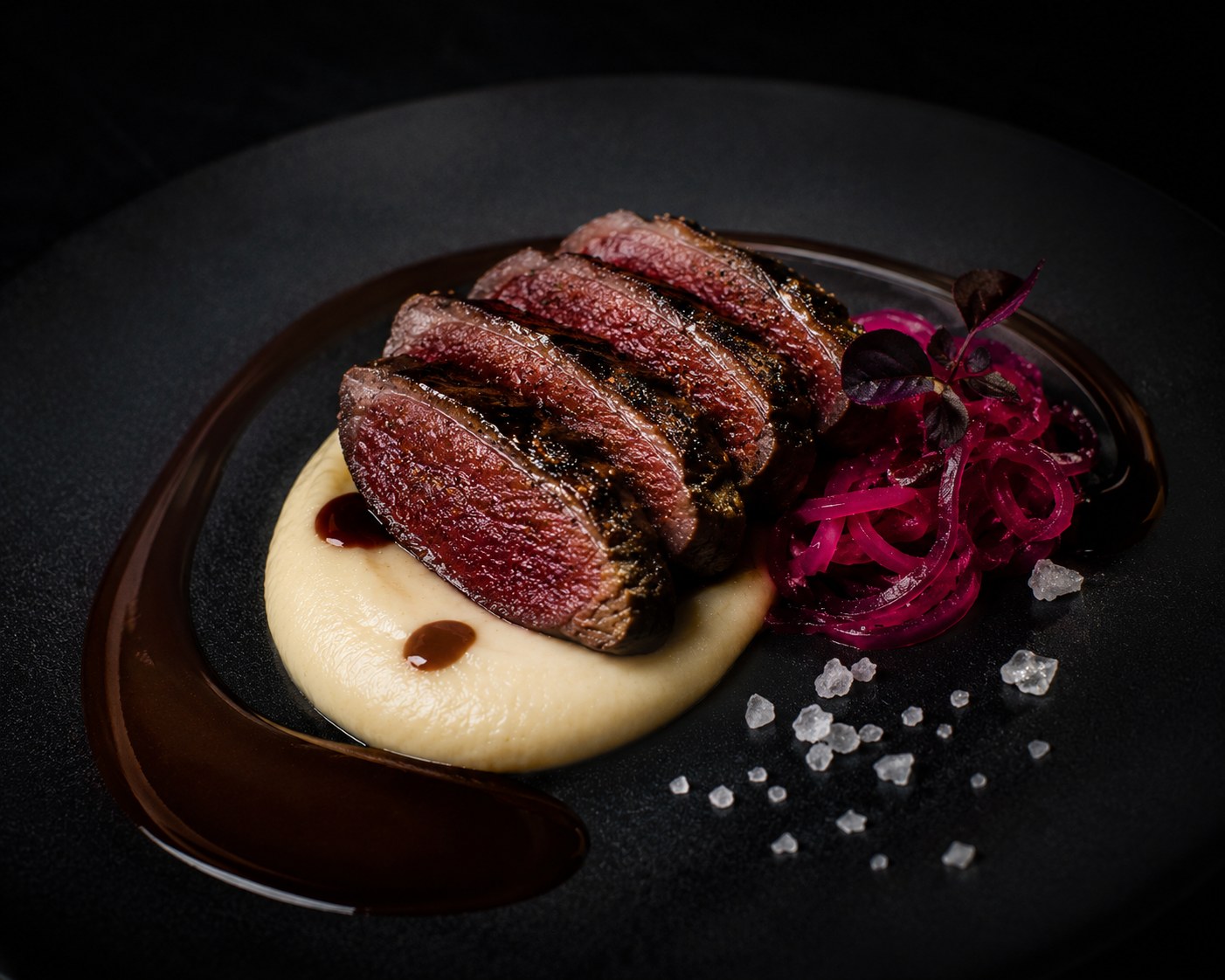 Pechugas de paloma zurita a la brasa rosadas en el corte sobre puré de chirivía ahumada con salsa oscura de entrañas y chocolate y cebolla encurtida en hibisco en plato negro