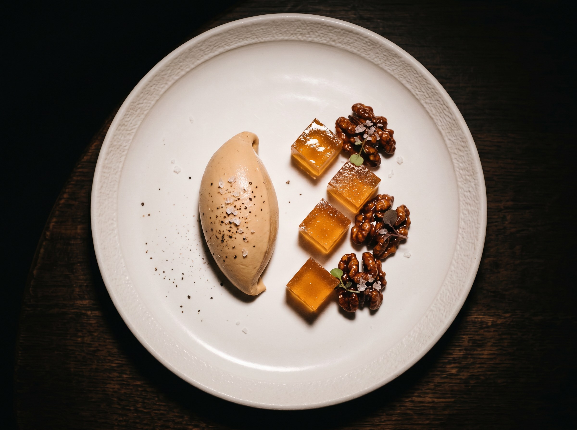 Hero final cenital: quenelle de parfait de foie gras con cubos de gelatina tawny y nueces caramelizadas alternados en diagonal, fondo negro