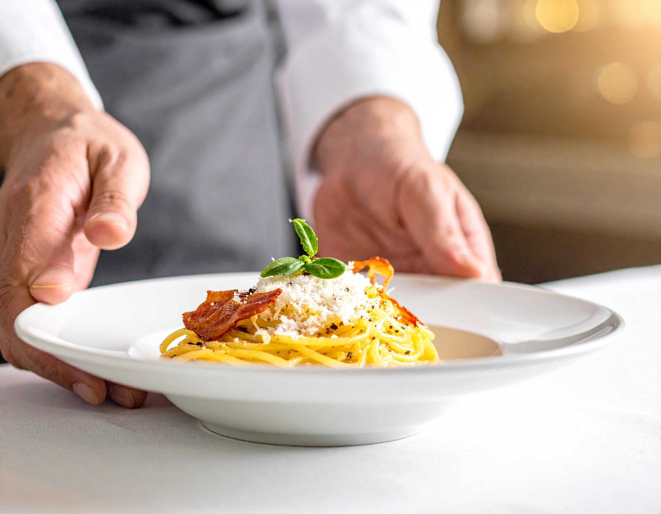 Chef de guantes blancos presentando plato de spaghetti carbonara con nevada de Pecorino Romano, guanciale crujiente y albahaca