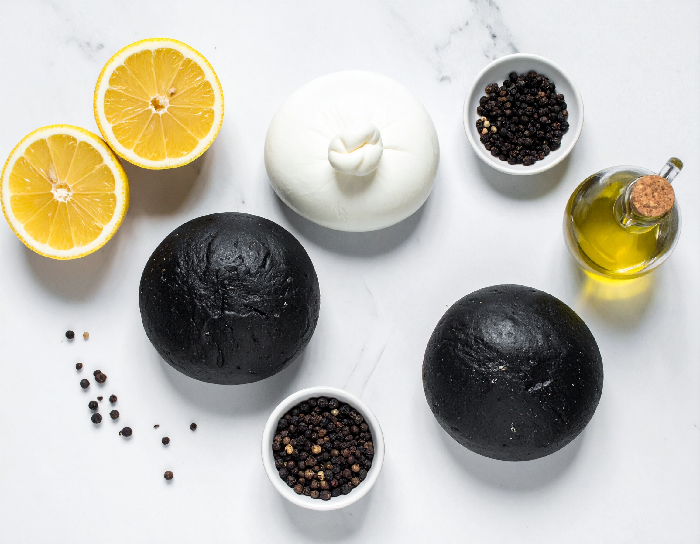 Flat lay ingredientes: masa negra, burrata, limón, aceite de oliva y pimienta sobre mármol blanco
