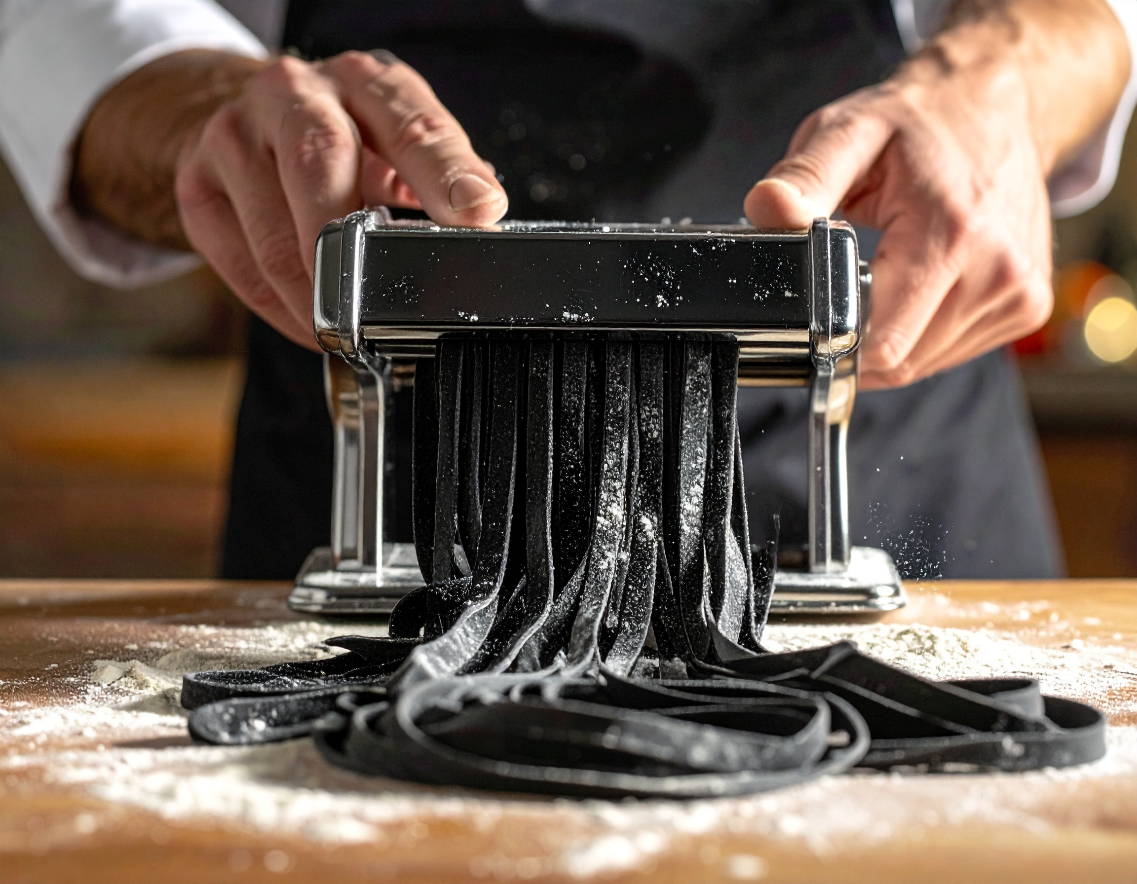 Tagliolini negros saliendo de la máquina de pasta, manos del chef guiando los fideos