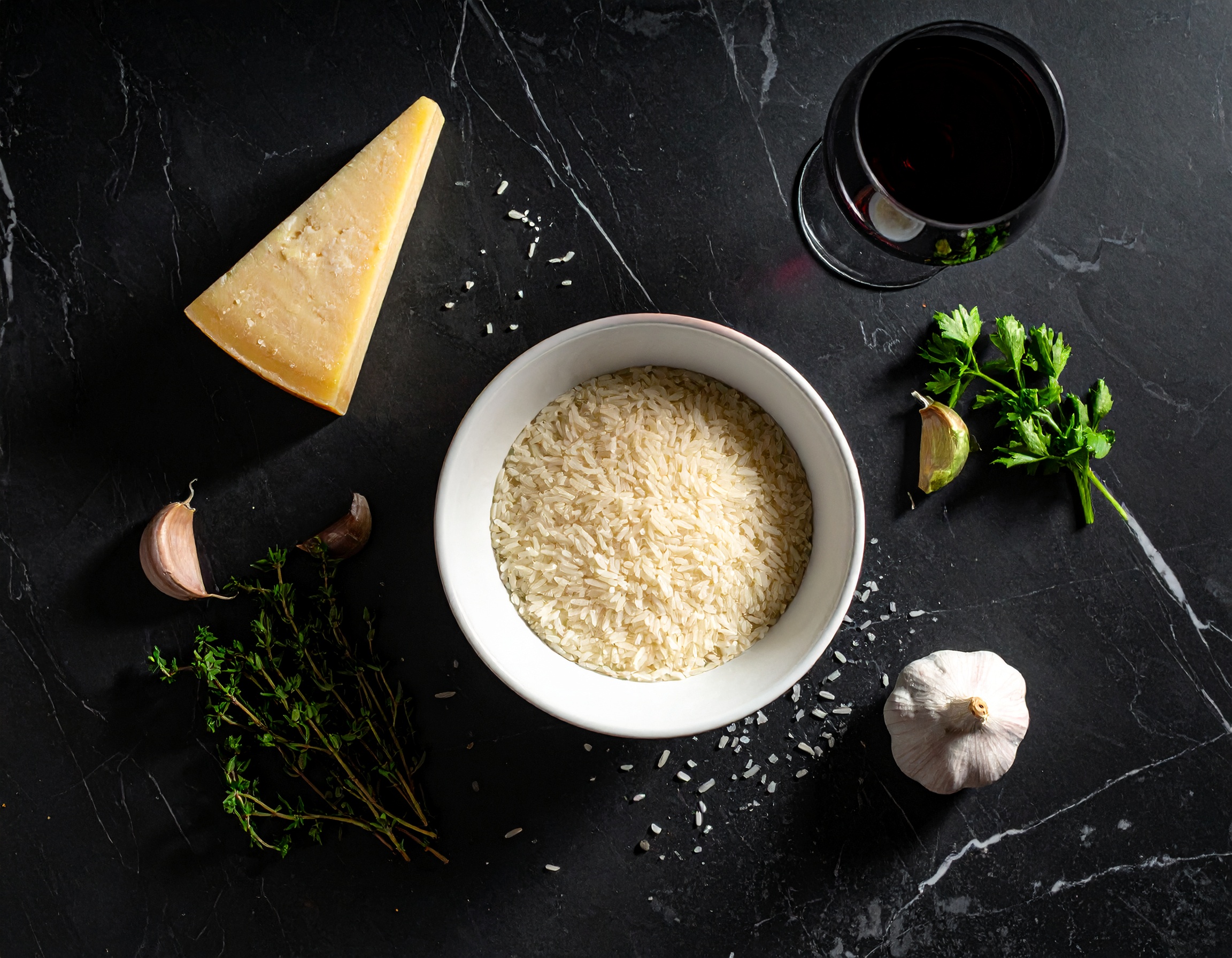 Flat lay de ingredientes del risotto negro sobre mármol oscuro: arroz arborio, cuenco de tinta, parmesano, mantequilla, vino blanco y hierbas