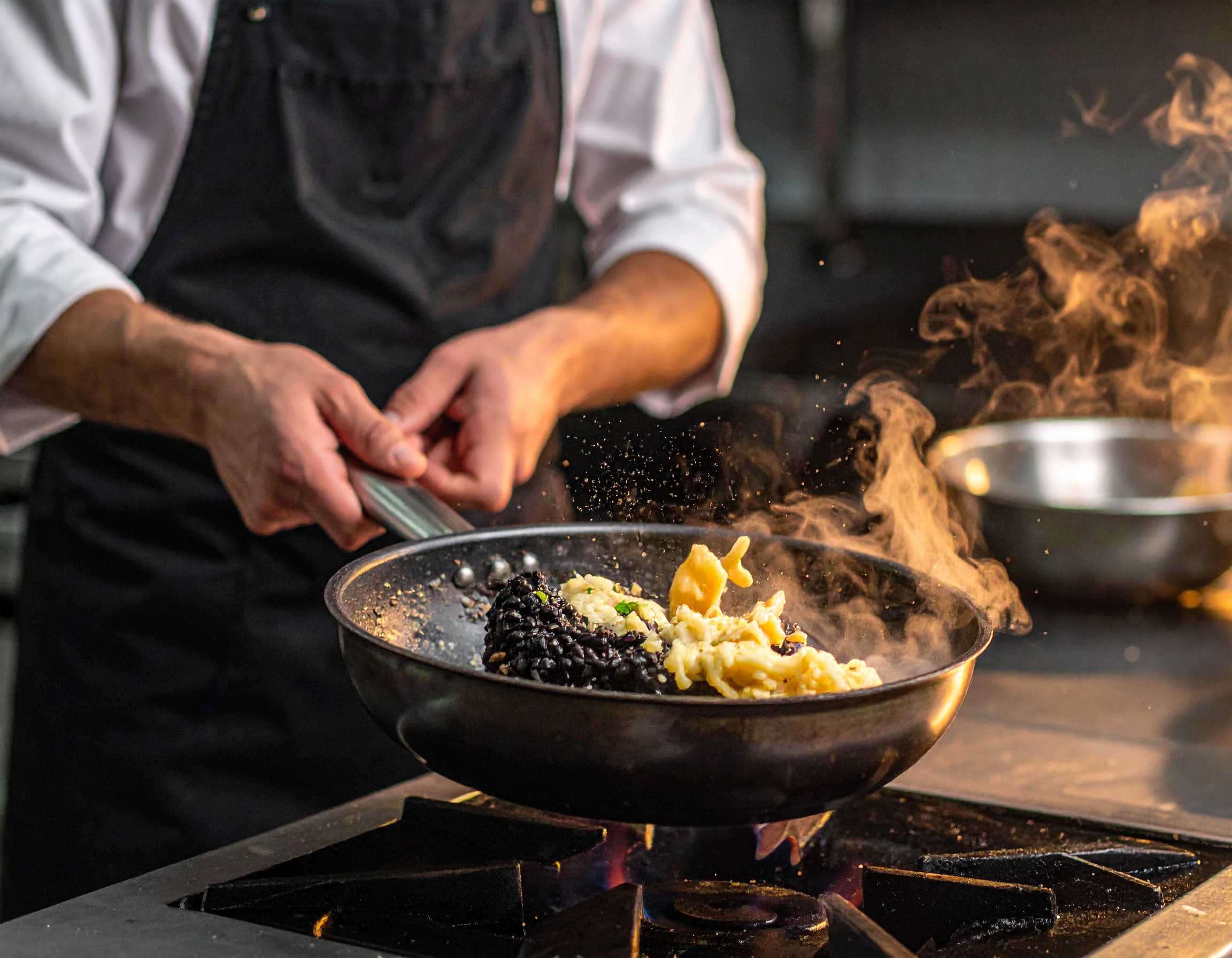 Mantecatura del risotto negro: chef agitando la cazuela fuera del fuego, ola cremosa de arroz negro con vapor