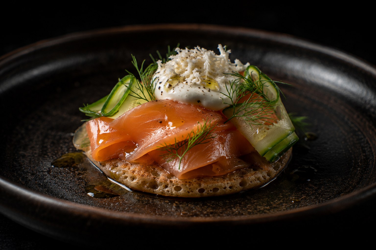 Láminas de trucha de río ahumada en frío sobre blinis de trigo sarraceno con crema de rábano picante fresco y pepino encurtido en dill sobre pizarra negra