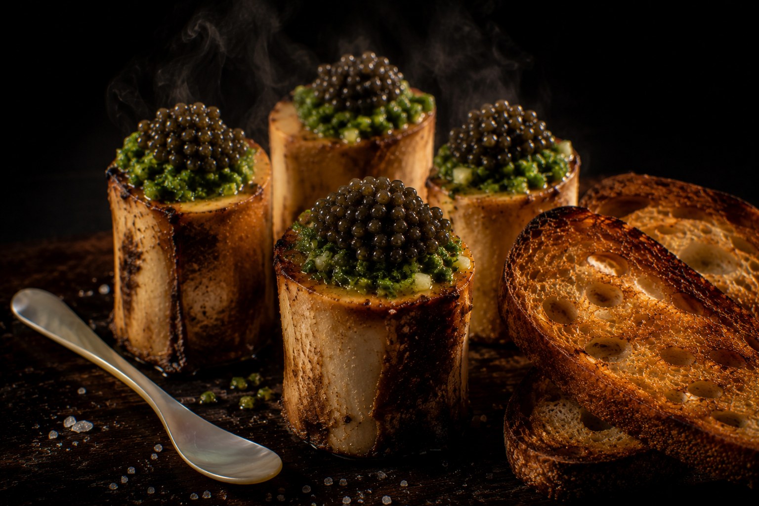 Huesos de tuétano de buey asados verticalmente con gremolata de yuzu verde brillante, perlas de caviar Osetra y pan de masa madre tostado en tabla de madera oscura